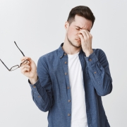 Guy rubbing eyes as feeling tired after wearing glasses while working over freelance project on computer standing with eyewear in hand and closed eyes being drained and exhausted, being hardworking