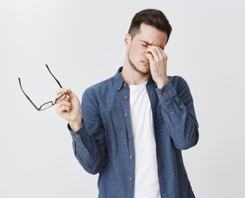 Guy rubbing eyes as feeling tired after wearing glasses while working over freelance project on computer standing with eyewear in hand and closed eyes being drained and exhausted, being hardworking