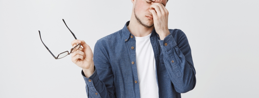 Guy rubbing eyes as feeling tired after wearing glasses while working over freelance project on computer standing with eyewear in hand and closed eyes being drained and exhausted, being hardworking