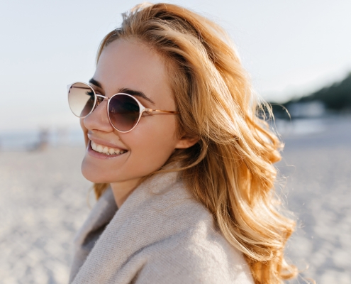 Close-up portrait of positive girl with wavy blonde hair dressed in beige cashmere sweater and sunglasses on beach.