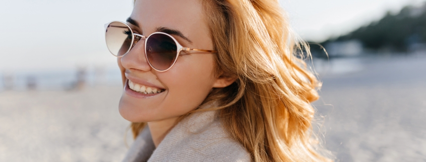 Close-up portrait of positive girl with wavy blonde hair dressed in beige cashmere sweater and sunglasses on beach.