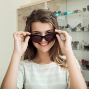 Pretty brunette girl smiling, trying on sunglasses in optical shop, asking boyfriend if they suit her, trying to find perfect eyewear before going on vacation to sea. Girl in store enjoys shopping