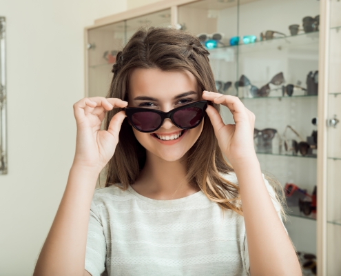 Pretty brunette girl smiling, trying on sunglasses in optical shop, asking boyfriend if they suit her, trying to find perfect eyewear before going on vacation to sea. Girl in store enjoys shopping